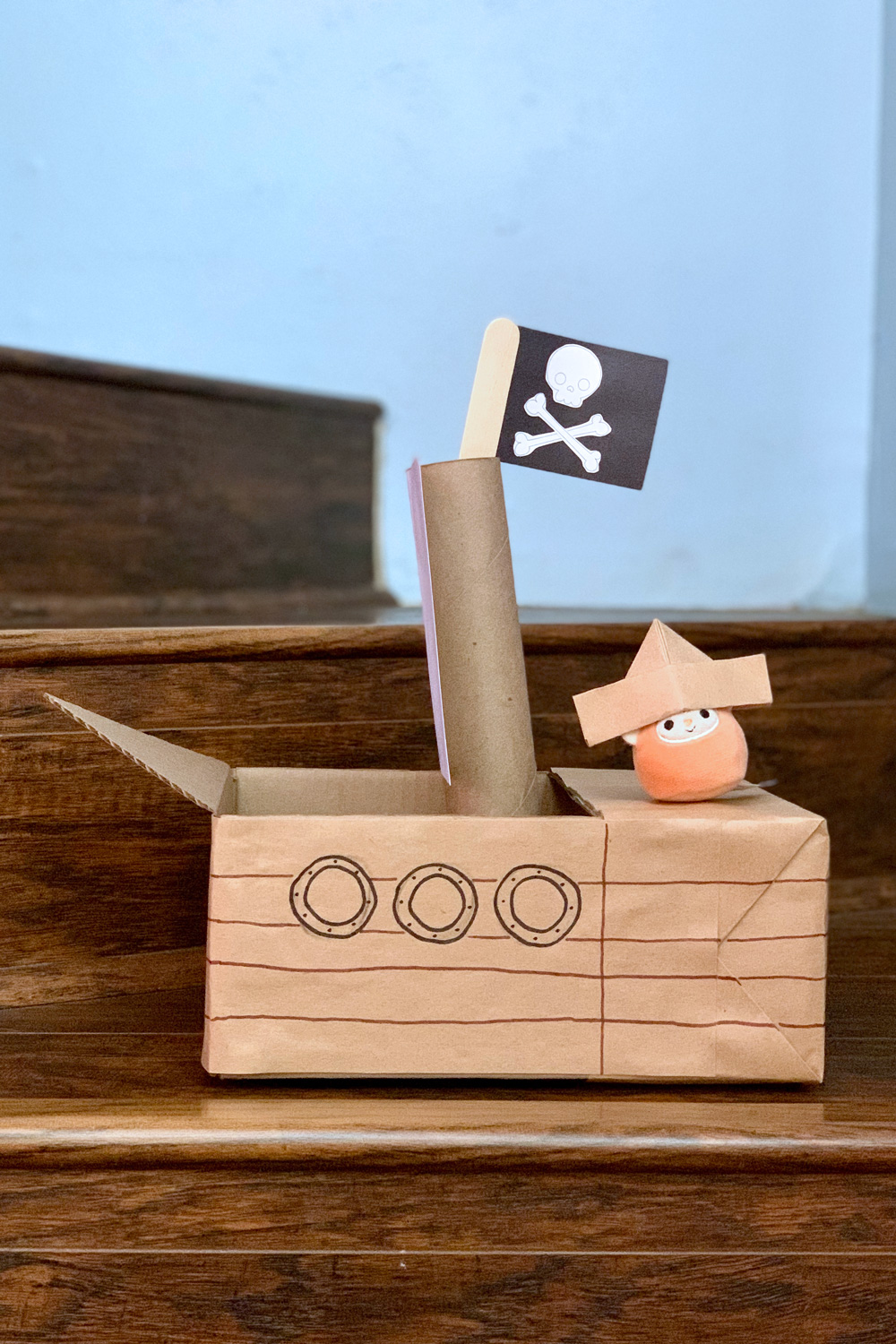 A cardboard pirate ship made from a box and paper tube with a pirate flag flying and a plushie wearing a paper hat.