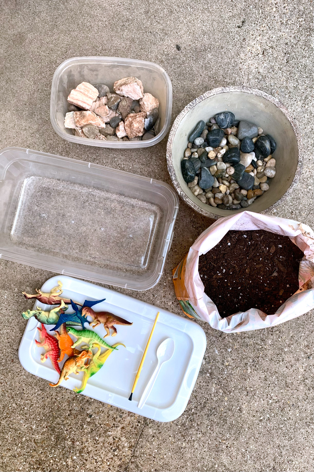 A box of fossils and rocks, a bag of potting soil, a clear plastic tub, toy dinosaurs, a plastic spoon, and a paintbrush.