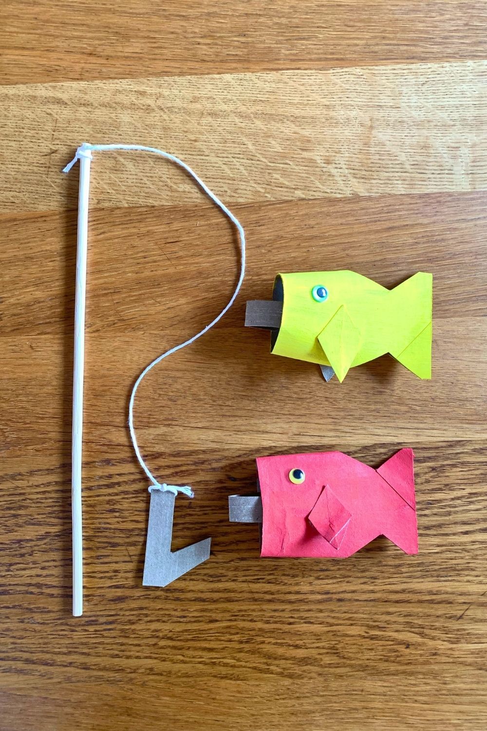 Pretend play fishing pole made from a dowel, string, and cardboard beside 2 paper fish.