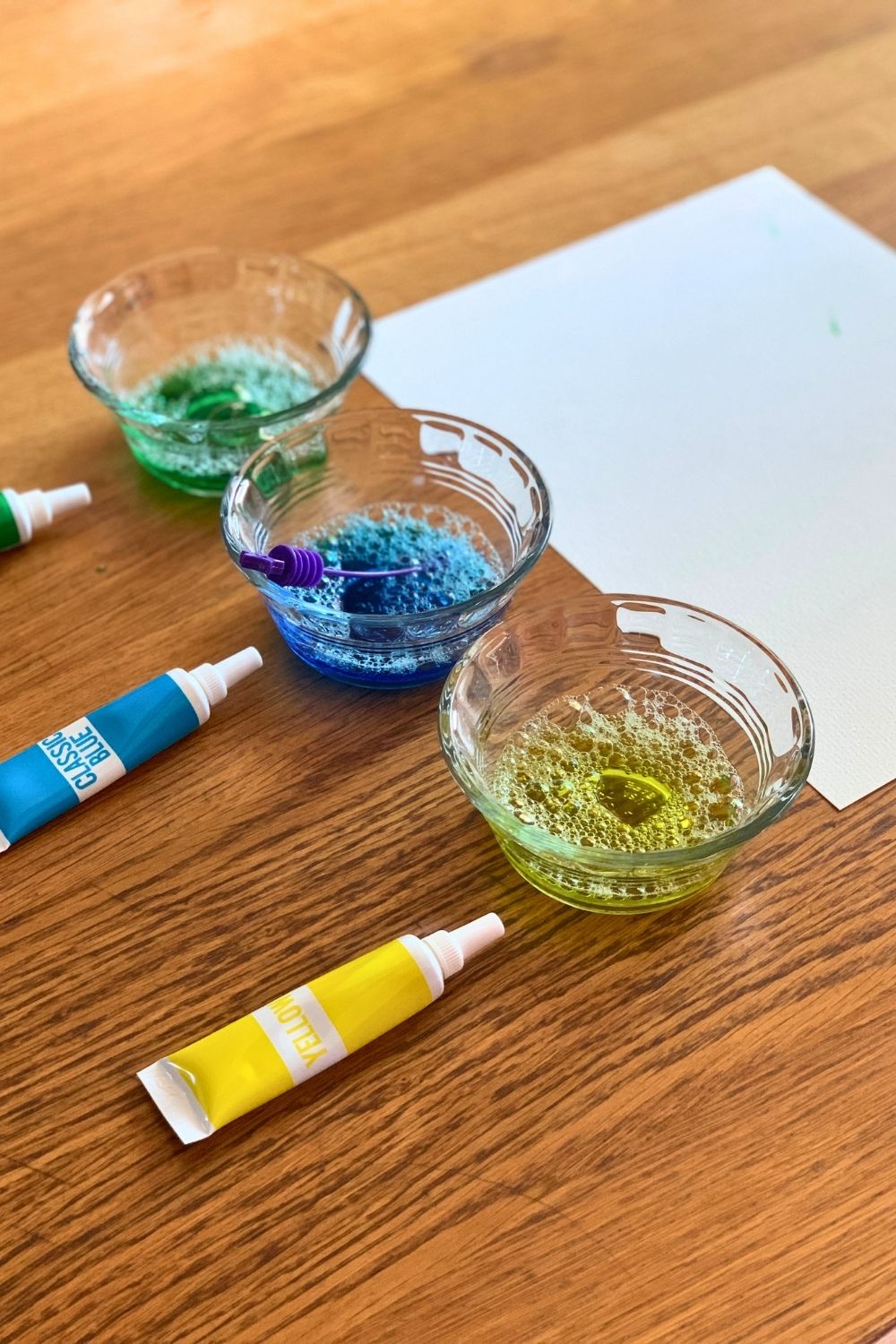 Three cups of bubbly colored water beside tubes of green, blue, and yellow food dye.