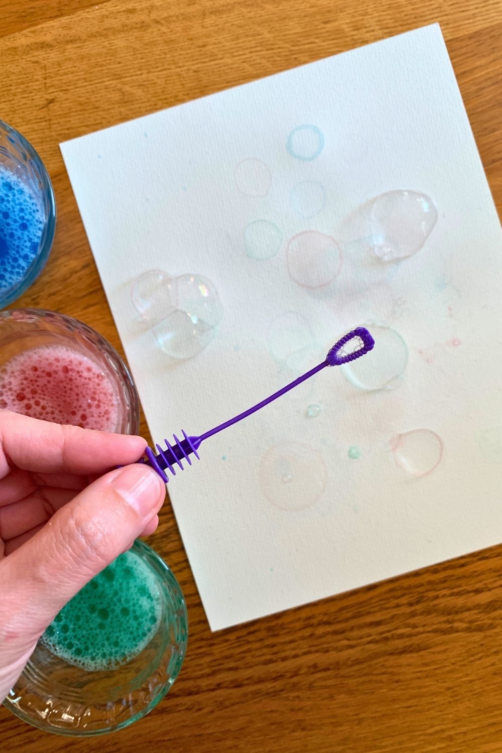 A hand holding a bubble wand over watercolor paper beside three cups of colored water.