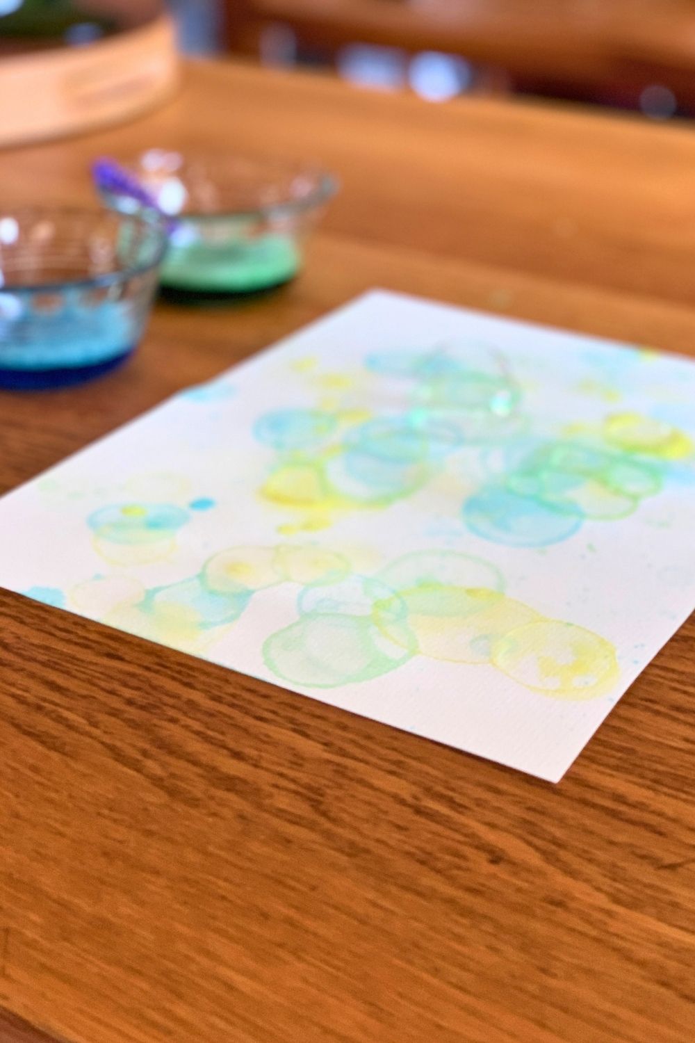 Vibrant blue, yellow, and green circles on watercolor paper beside cups of mixed bubble paint.