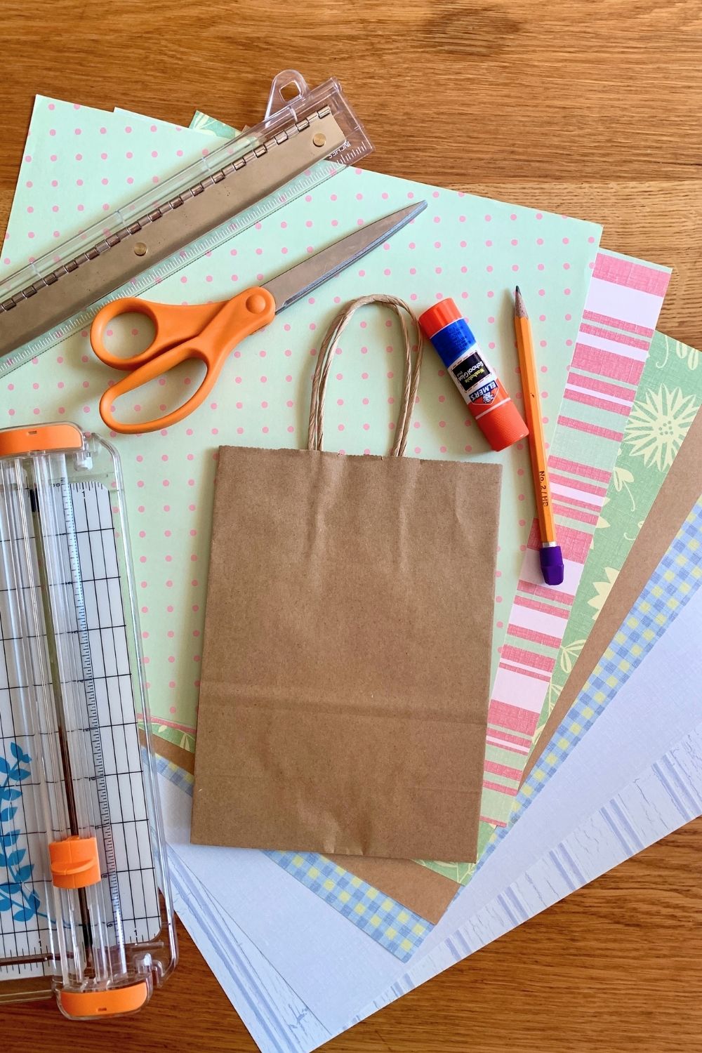 A brown kraft paper gift bag on a stack of colorful scrapbook paper, a ruler, scissors, gluestick, pencil, and paper trimmer.
