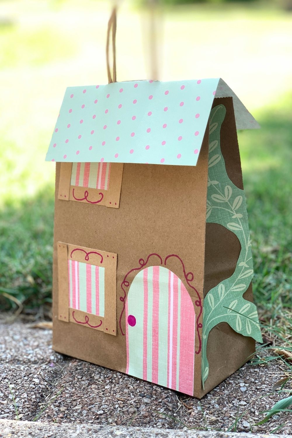 A paper bag fairy house from a brown gift bag decorated with scrapbook paper roof, doors, windows, and plants.