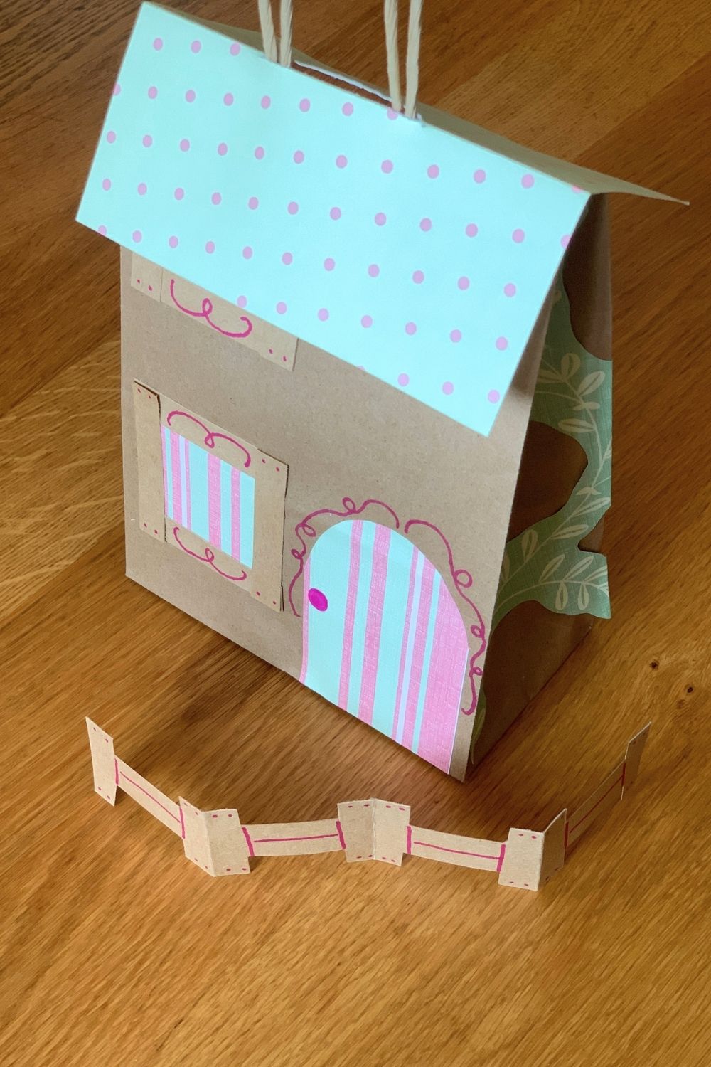 A paper bag play fairy house with a triangle roof, door, windows, and a little fence.