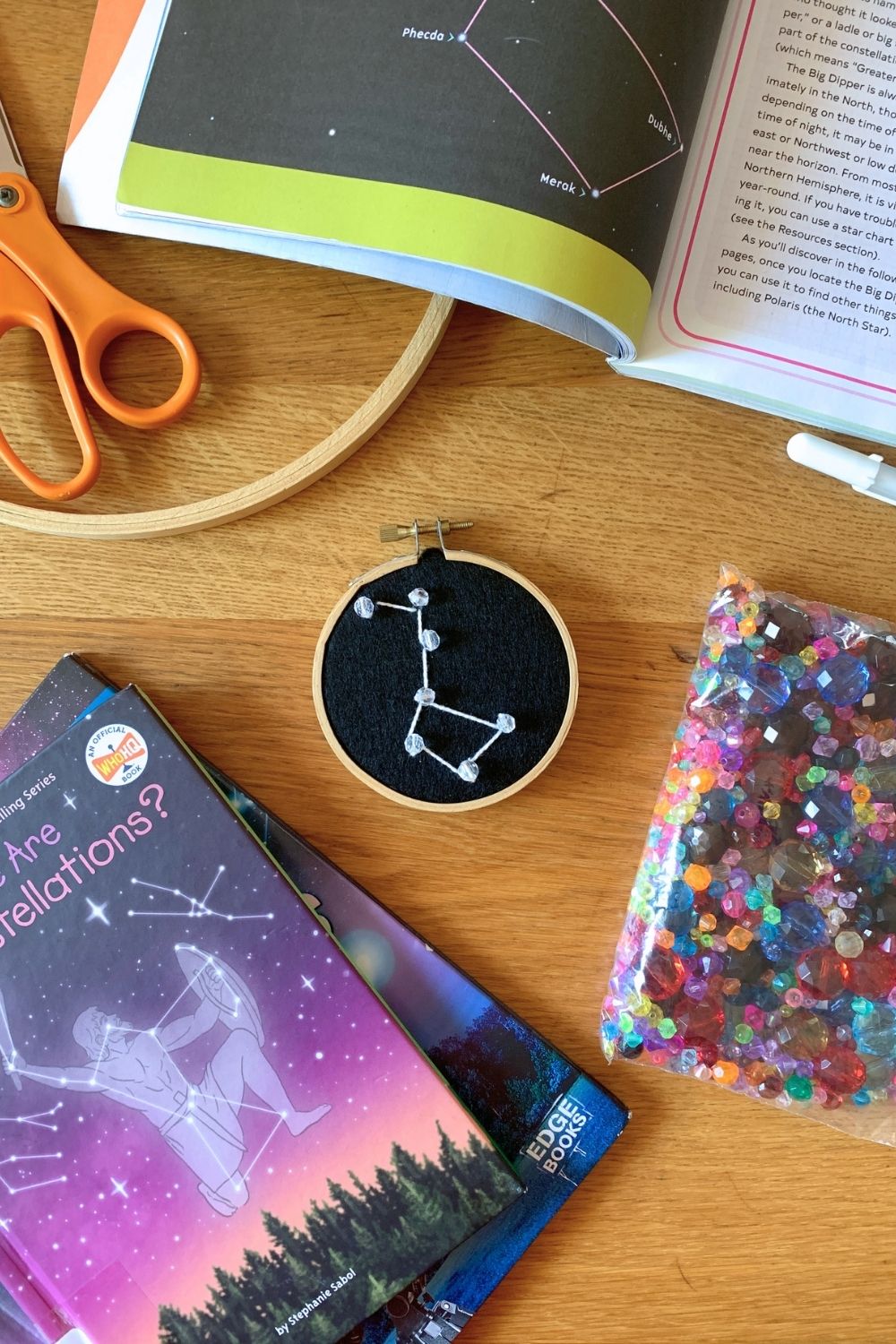 Embroidery of the big dipper constellation in white on a black background surrounded by books and embroidery supplies.