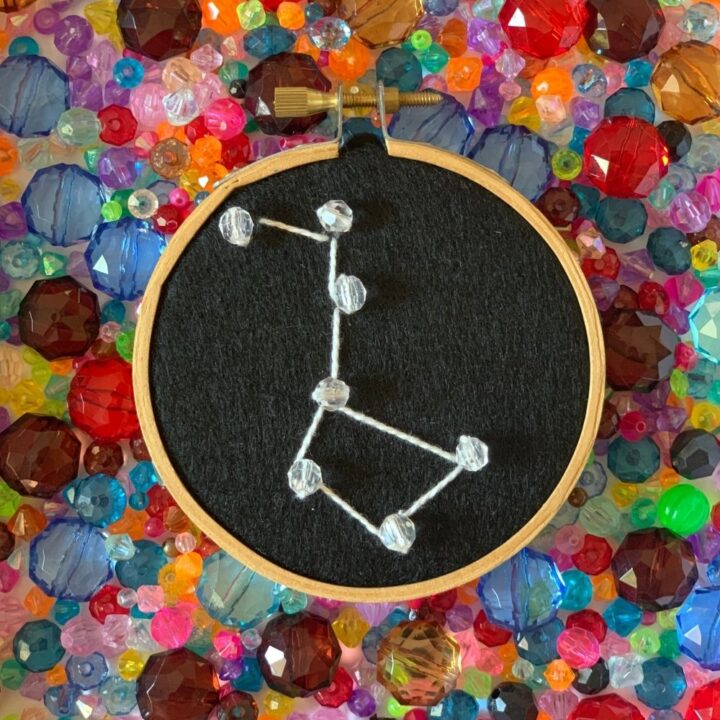 An embroidery hoop with the big dipper constellation in white thread on a black background, beads at the stars.