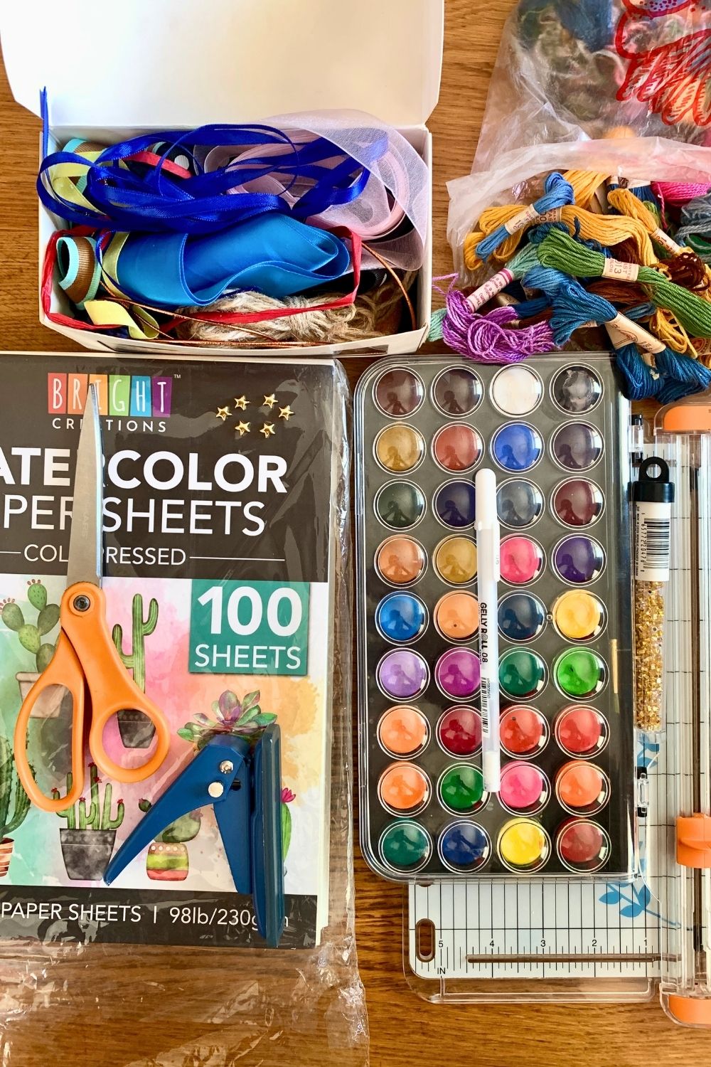 Watercolor paper and paint palette beside a box of ribbon, bag of embroidery thread, paper trimmer, scissors, hole punch, white gel pen, and beads.